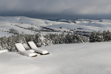 Two wooden sun loungers covered with snow on the top of the hill in winter.