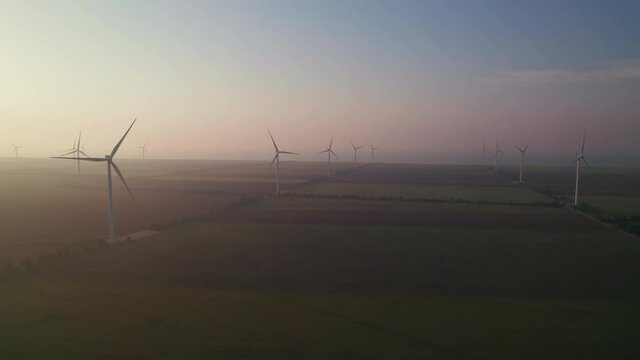 Windmills with rotating wings at sunrise or sunset or sunrise. Technology for wind energy, renewable energy source, earth care. Wind power station, drone video, moving toward the horizon, 4k