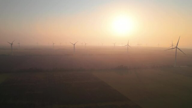 Windmills with rotating wings at sunrise or sunset or sunrise. Technology for wind energy, renewable energy source, earth care. Wind power station, drone video, moving toward the horizon, 4k