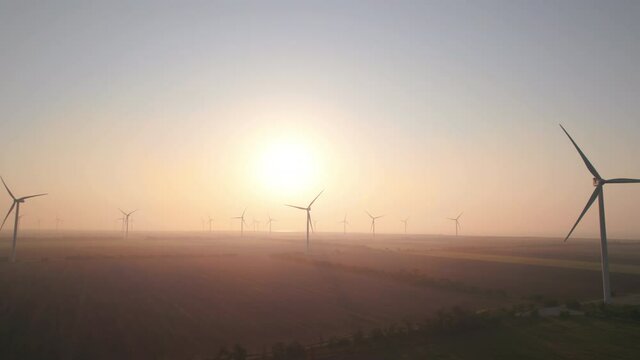 Windmills with rotating wings at sunrise or sunset or sunrise. Technology for wind energy, renewable energy source, earth care. Wind power station, drone video, moving toward the horizon, 4k