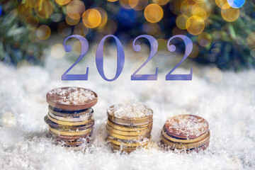 Date 2022. Gold pyramids of coins covered with snow on a background of green spruce branches and bokeh from a garland