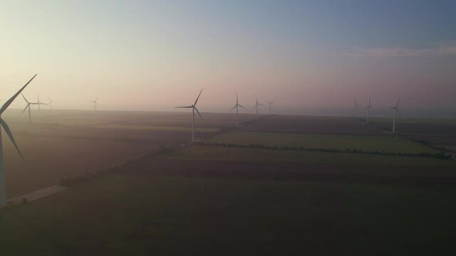 Windmills with rotating wings at sunrise or sunset or sunrise. Technology for wind energy, renewable energy source, earth care. Wind power station, drone video, moving toward the horizon, 4k