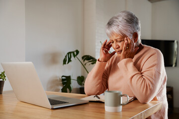 Multi-ethnic elderly female holding her head in pain. Sitting with headache at modern kitchen counter at home.