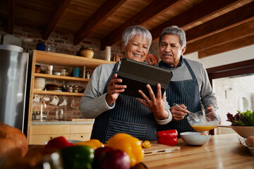 Happy Multi-cultural elderly couple smiling, using electronic tablet to research recipe in modern kitchen.