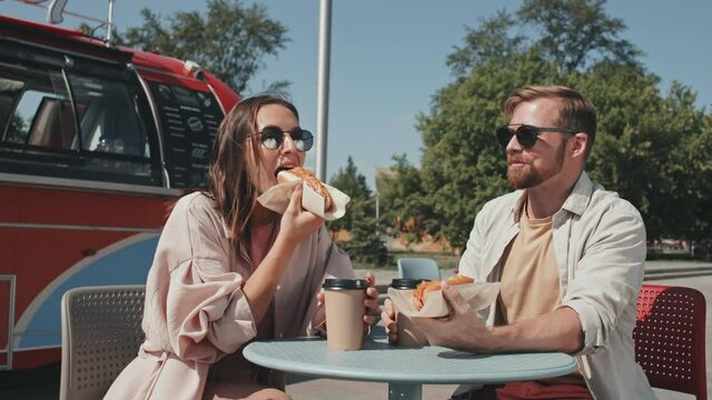 Medium shot of young Caucasian couple eating hotdogs with coffee and having conversation sitting at table next to food truck on sunny day