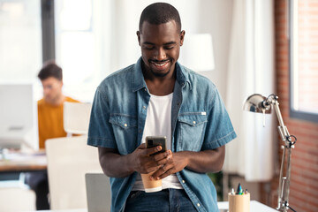 Happy businessman using smartphone while drinking a cup of coffee sitting on a desk in a coworking space.