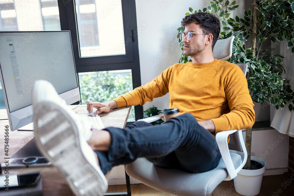 Poster graphic designer working with computer while drinking coffee with feet on the desk in modern startup