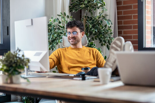 Graphic designer working with computer while drinking coffee with feet on the desk in modern startup office.