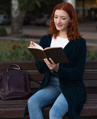 Obraz premium A young and attractive redhead Caucasian girl is reading a book while sitting on a bench. Education concept.