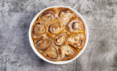 Cinnamon rolls or cinnabon, homemade sweet traditional dessert buns with white cream sauce in a white baking dish on a dark grey background. Top view, flat lay