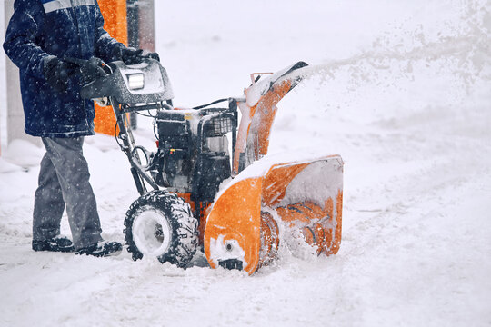 Snow blower machine during snowfall. Worker blowing snow during blizzard. Man using snowblower, clearing snow from road at parking lot.