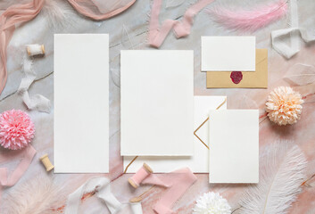 Wedding cards and envelopes between pastel flowers, silk ribbons and feathers on marble