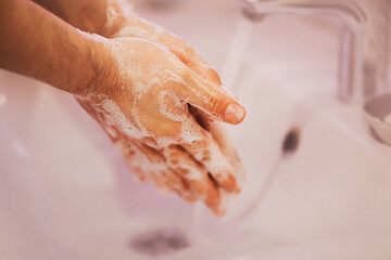 Fototapeta premium A man carefully washes his hands with soap suds over a white ceramic sink in the bathtub. Cleanliness and hygiene.
