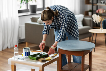 renovation, diy and home improvement concept - woman in gloves with paint brush painting old wooden table to grey color