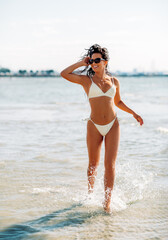 people, summer and swimwear concept - happy smiling young woman in bikini swimsuit running in water beach