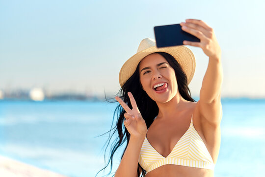 people, summer and swimwear concept - happy smiling young woman in bikini swimsuit and straw hat taking selfie with smartphone on beach