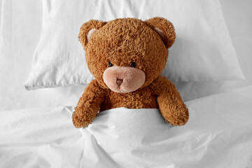 bedtime, sleeping and childhood concept - teddy bear toy lying on pillow covered with blanket in bed