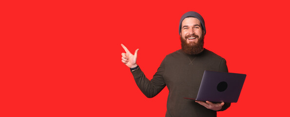 Smiling male with netbook pointing away while standing against red background and looking at camera