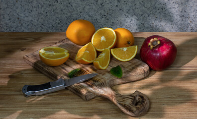 Still life: a group of fresh oranges, whole and cut, lies on an old figured wooden board, and next to it is a ripe pomegranate and a knife.  Drawing with light and shadow. Selective focus