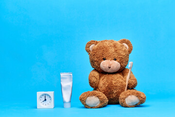 dental care, health and childhood concept - brown teddy bear with toothbrush, toothpaste and alarm clock over blue background