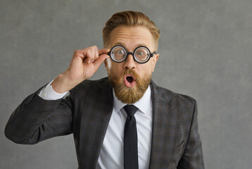 Fototapeta na wymiar Portrait of shocked and funny caucasian businessman in glasses with magnifying glass. Man dressed in a stylish costume on a gray concrete background. Concept of considering new business ideas.