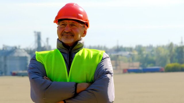 Grain storage modern technology. Positive manager in orange hardhat crosses arms looking at installed equipment in field against large silo
