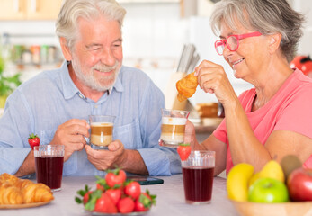 Cheerful caucasian couple having breakfast at home with milk, coffee, croissant. Senior people relaxed and happy, enjoying food and drink