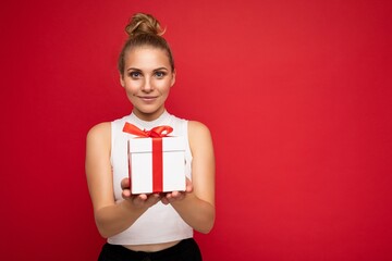 Photo shot of beautiful attractive cute positive young blonde woman isolated over red background wall with copy space for text wearing casual white top holding gift box with red ribbon. Birthday