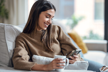 Smiling young woman using her mobile phone while drinking coffee sitting on sofa at home.