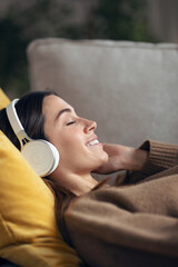 Beautiful woman listening to music with headphone while using smartphone lying on sofa at home.