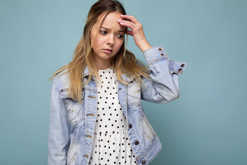 Photo of young sad upset attractive blonde woman with sincere emotions wearing jean blue jacket isolated over blue background with free space and having headache