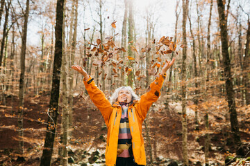 Senior woman, in a forest, happily throwing brown tree leaves to the air in autumn