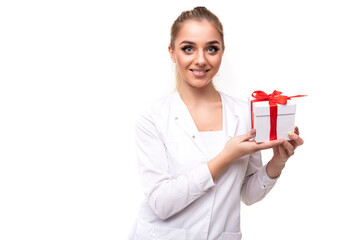 Photo of a beautiful pretty attractive positive smiling young blonde woman with a ponytail and makeup in a white medical coat of a nurse holding a white gift box with a red ribbon isolated on a white
