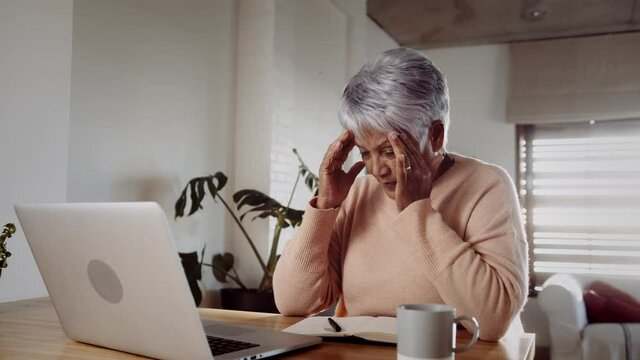 Multi-cultural elderly female upset, recieving bad news while on laptop, sitting at kitchen counter