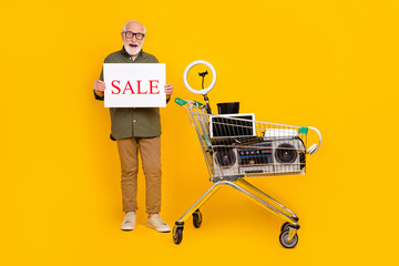 Full size portrait of positive aged man hold sale banner old stuff items trolley isolated on yellow...