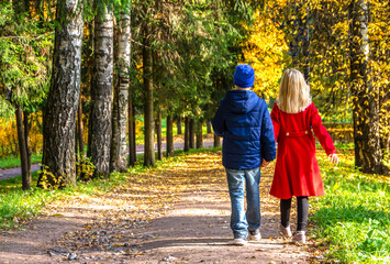 Fototapeta premium A blonde girl and a boy holding each other's hands (a couple) walk along the road in an autumn park