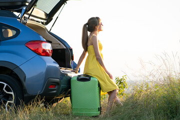 Young female driver having rest sitting on a green suitcase near her car in summer nature. Travel and vacations concept.