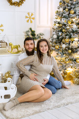 A young couple near the Christmas tree. A young pregnant girl. A young guy with a beard.