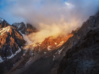 Sunlight in the mountains. Big glacier on top in orange light. Scenic mountain landscape with great snowy mountain range lit by dawn sun among low clouds.