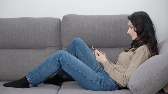 Woman browsing internet on mobile phone while lying on comfortable couch in living room at home. Stock video clip of beautiful middle aged female model using modern smartphone to read news