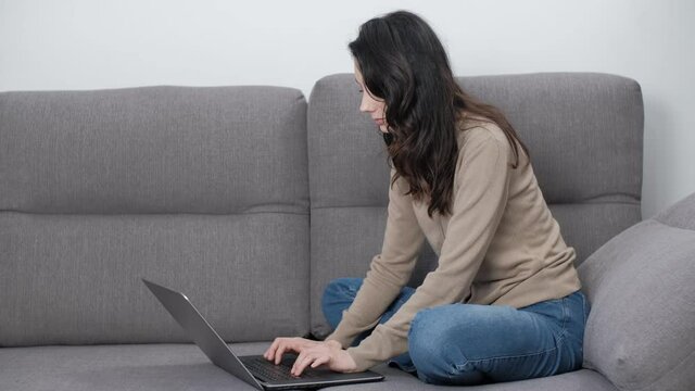 Stock video of freelancer woman working on notebook computer from home. Free lance worker female typing text on laptop keyboard while sitting on couch in living room