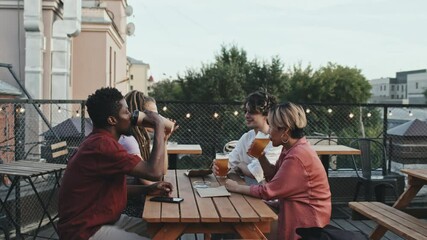 Medium shot of cheerful multiethnic male and female friends toasting with beer glasses sitting together at outdoor rooftop cafe decorated with yellow lights on summer evening - Powered by Adobe