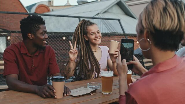 Medium shot of young woman taking photo of her female friend showing piece sign drinking beer and having good time together at outdoor rooftop cafe on summer evening