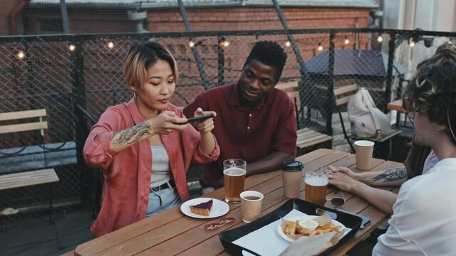 Medium shot of young Asian woman spending summer evening together with multiethnic friends, taking pictures of food on table, sitting at cozy rooftop bar with beer and snacks