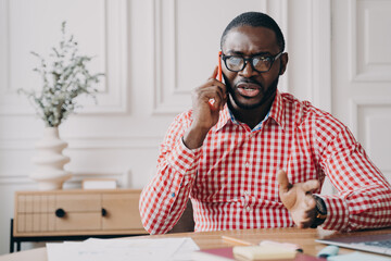 Dissatisfied African American businessman emotionally speaking on smartphone while sitting at desk