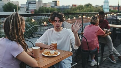 Medium shot of young hipster man with glass of beer talking to his girlfriend sitting together at table in outdoor rooftop bar on summer evening - Powered by Adobe