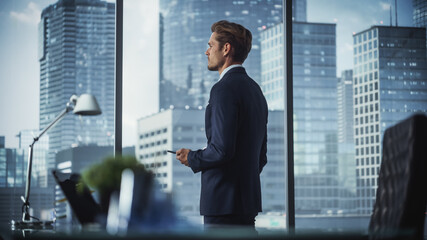 Fototapeta premium Successful Young Businessman in a Suit Standing in His Modern Office, Using Smartphone, Looking out of the Window on Big City with Skyscrapers. Successful Finance Manager Planning Work Projects.