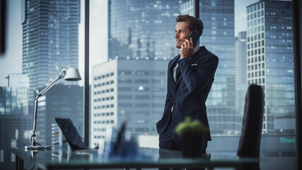 Confident Young Businessman in a Suit Standing in Modern Office, Talking on a Phone, Looking out of...