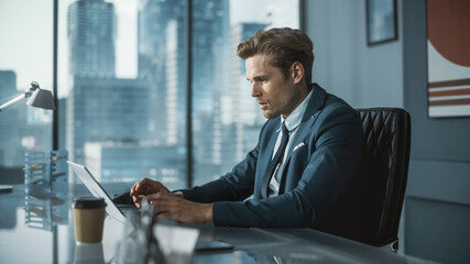 Confident Businessman in a Suit Sitting at a Desk in Modern Office, Using Laptop Computer, Next to Window with Big City with Skyscrapers View. Successful Finance Manager Planning Work Projects.