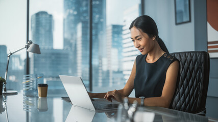Successful Businesswoman in Stylish Dress Sitting at a Desk in Modern Office, Using Laptop...